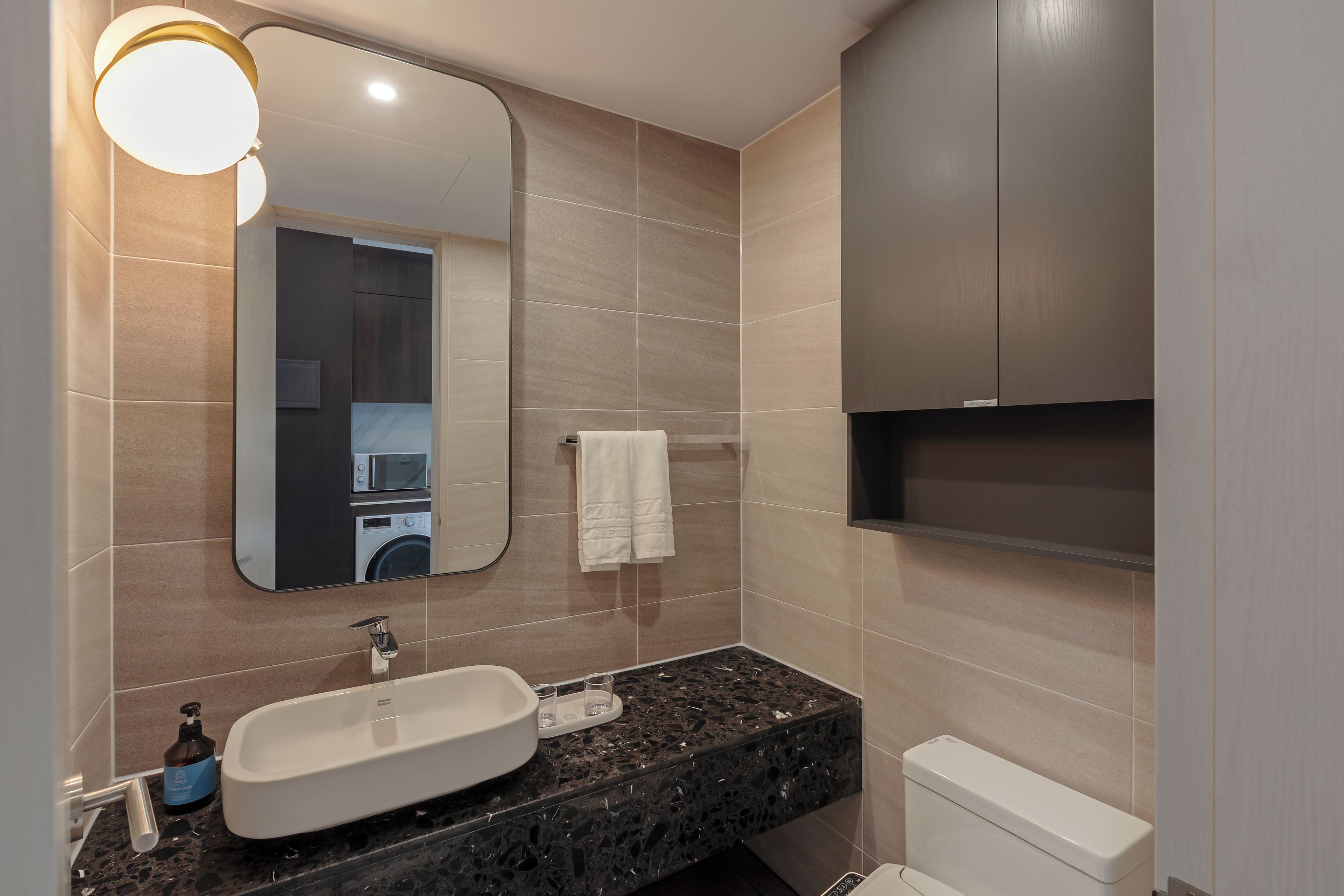A modern bathroom with a large mirror, a white sink, a black marble countertop, a toilet, and a towel hanging on the wall.
