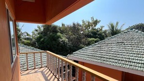 A wooden balcony with a railing, a window, and a view of a green-roofed building and trees. - Kawari Resort (Gokarna)