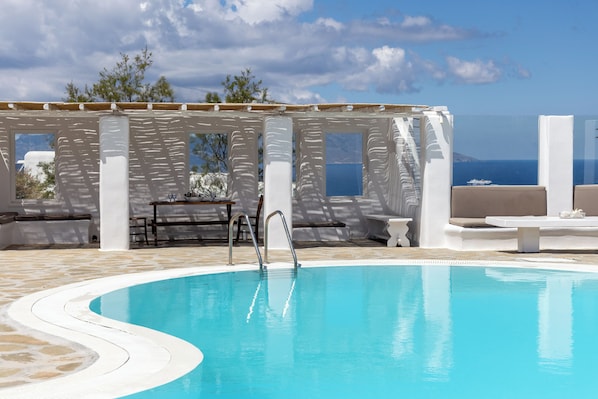 A pool area with a clear view of the sea, a pergola, and a seating area with a table and chairs.