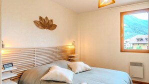 A bedroom with a bed, bedside tables, a wall-mounted lamp, a window with a view of houses, and a decorative leaf on the wall. - Apartment in Thones With Mountain Views (Thones)