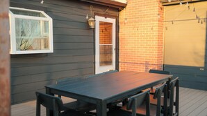 A wooden deck with a dark-colored table and chairs, a white-framed window, and a brick wall. - The Vinyl House: Olde Town - King Bed - EV Charger - Epic Views - Designer Home (Arvada)