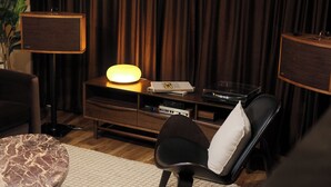 A room with a dark curtain backdrop, a table with a lamp, a chair with a pillow, and a speaker on a wooden stand. - The Vinyl House: Olde Town - King Bed - EV Charger - Epic Views - Designer Home (Arvada)