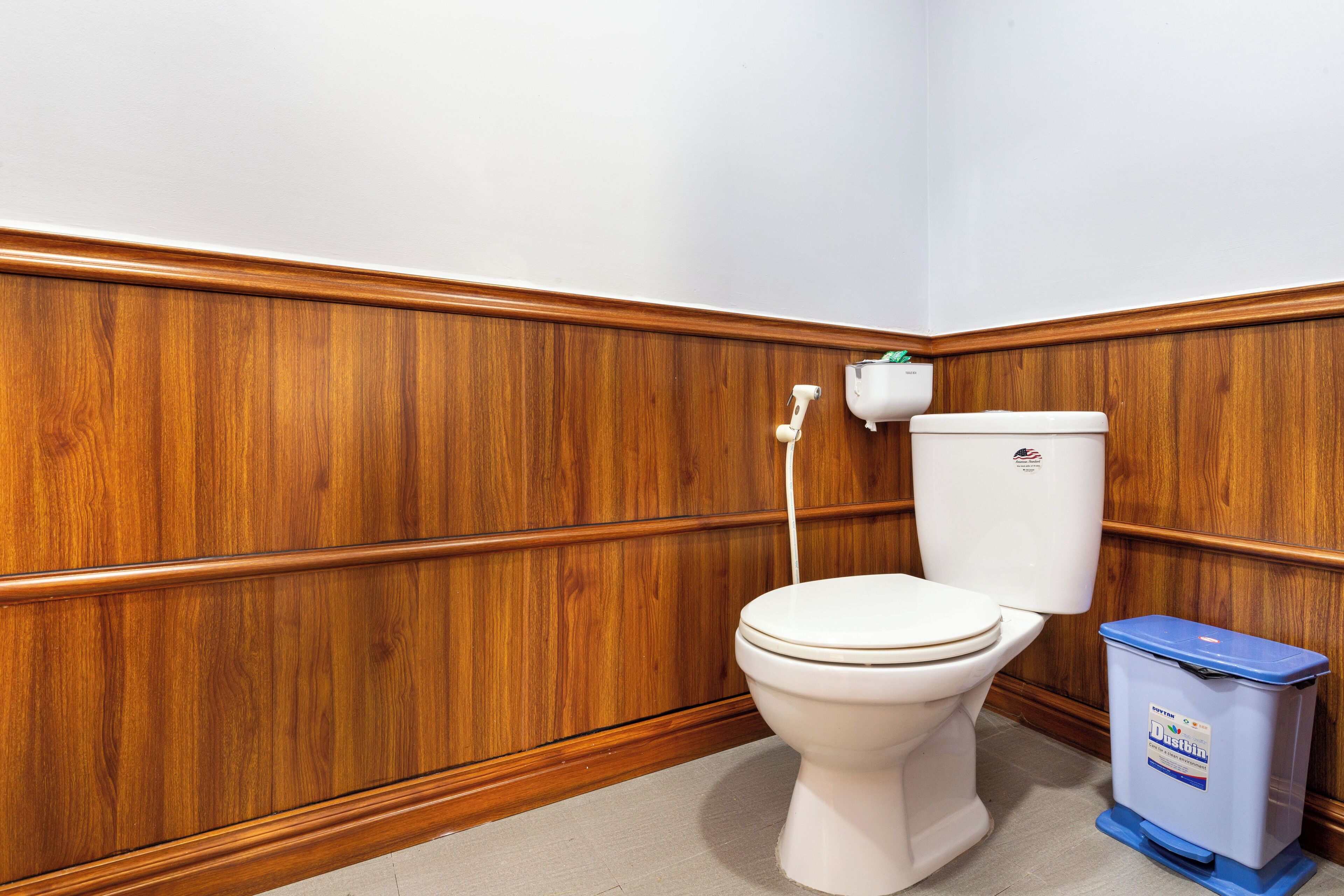 Une salle de bain avec des toilettes blanches, un lambris en bois et une poubelle bleue.