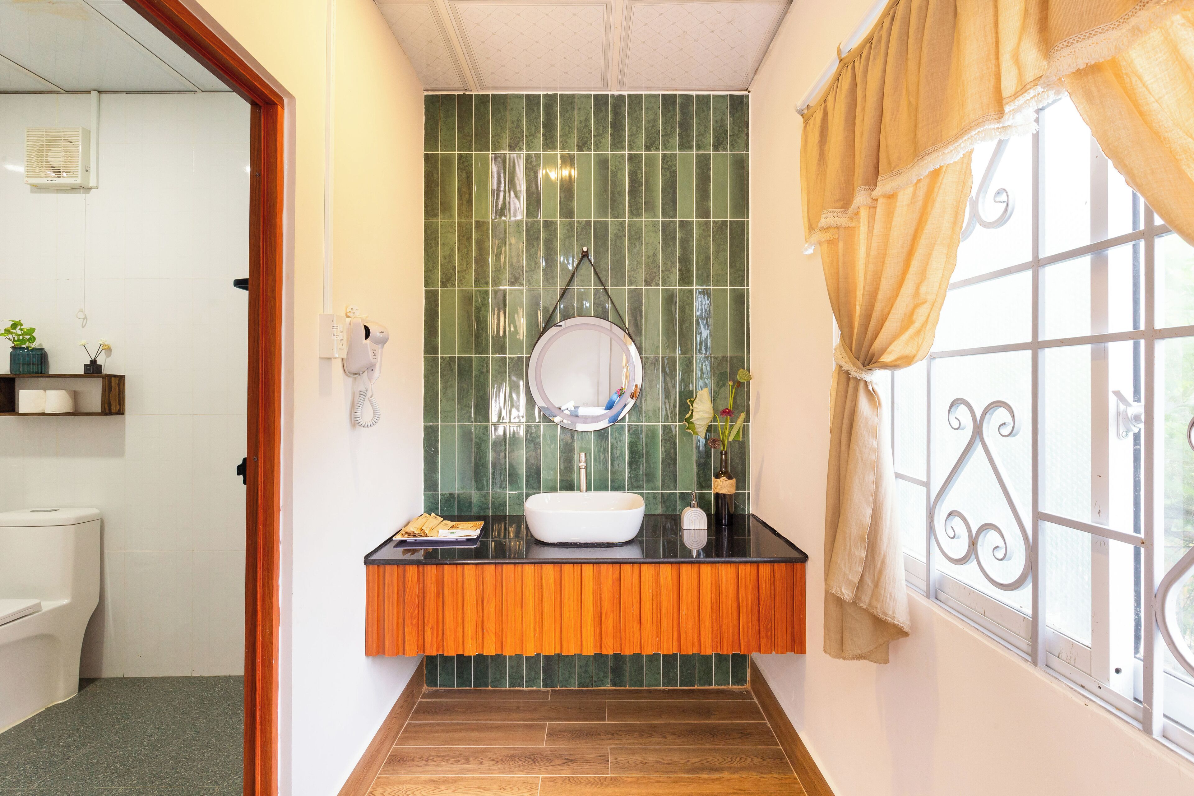 Un baño con un lavamanos blanco, salpicadero de azulejos verdes y una ventana con cortina.