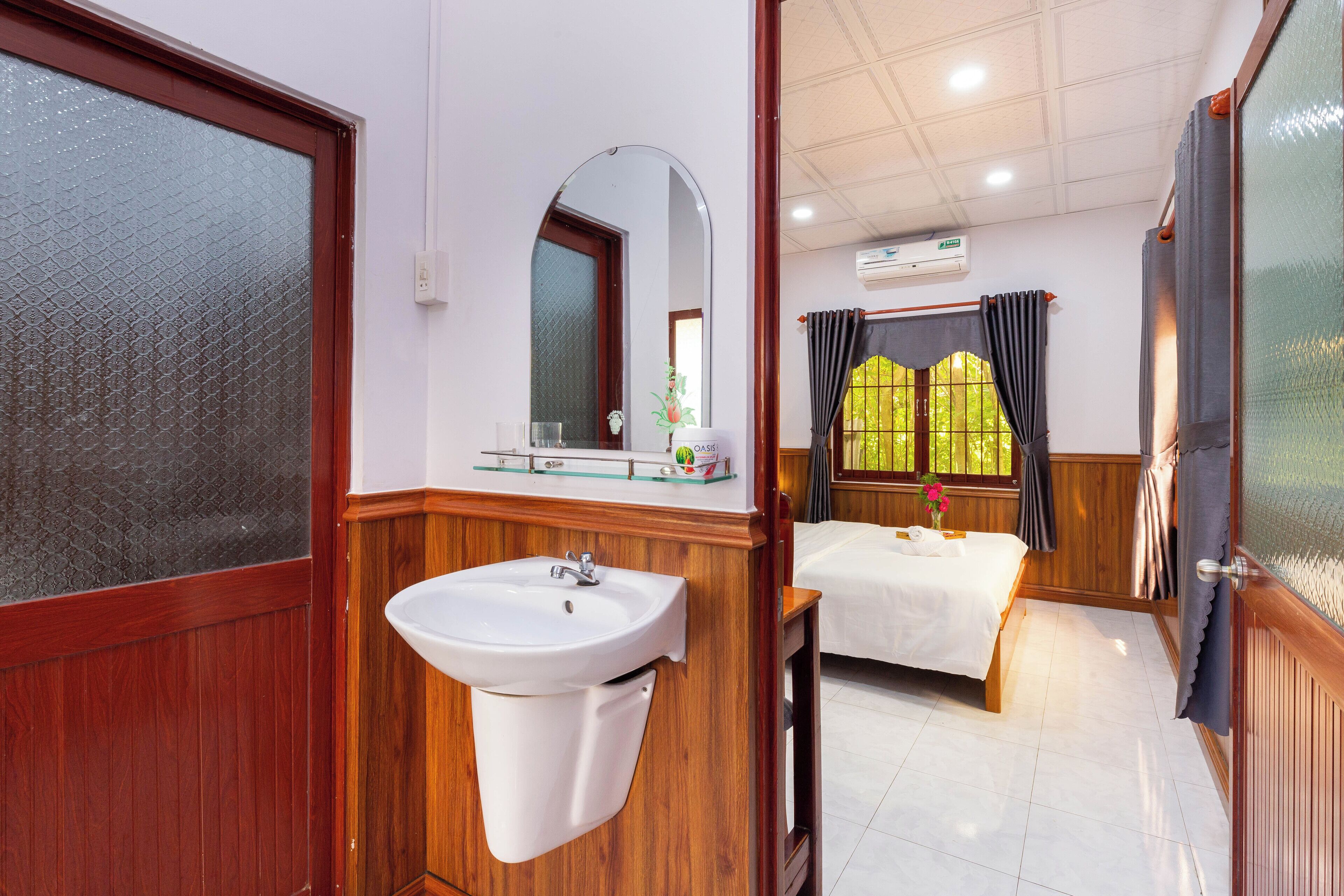 A hotel room with a wooden vanity, a bed with a white bedspread, a window with curtains, and a mirror above the vanity.