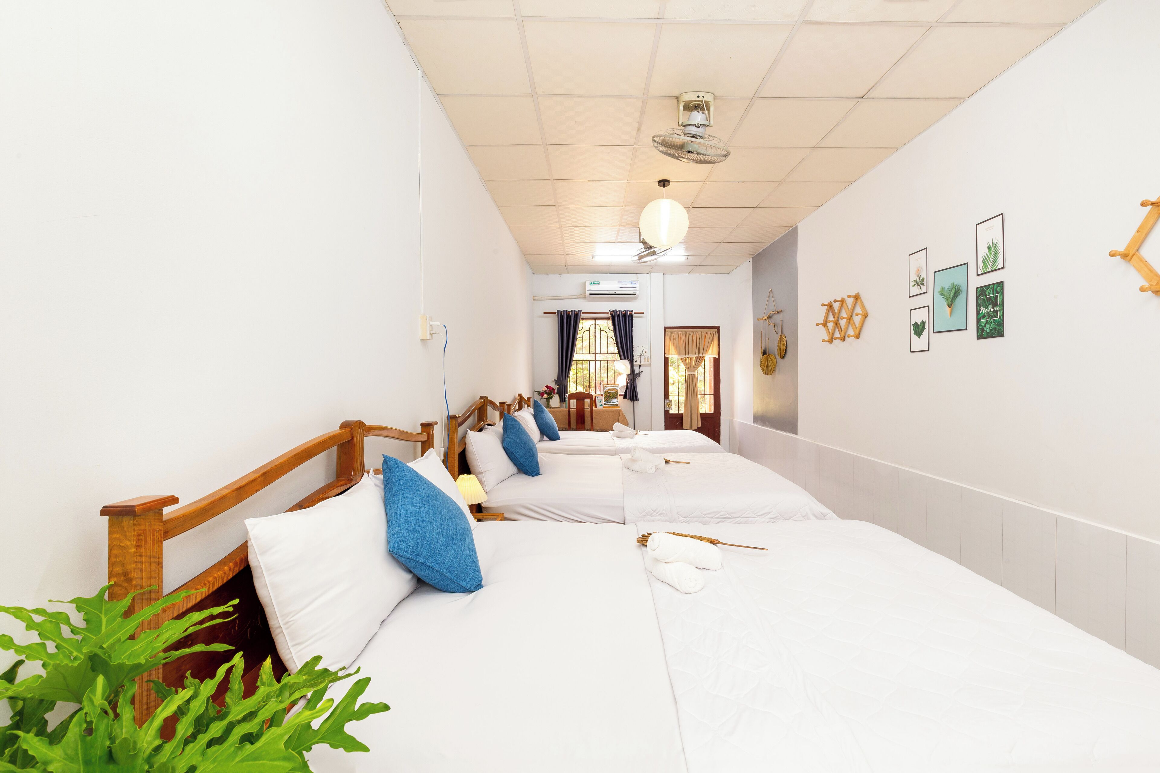 Ein Hotelzimmer mit drei Einzelbetten, einem Holzkopfende, einem Deckenventilator und einer Wandklimaanlage.