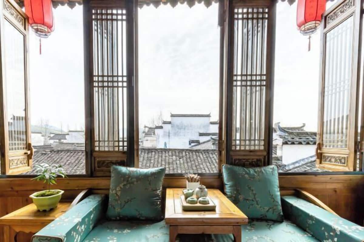 Une pièce de style traditionnel, avec un banc en bois, une petite table sur laquelle est disposé un service à thé, et une vue sur des bâtiments traditionnels, visible à travers une fenêtre ornée d’un motif en treillis.
