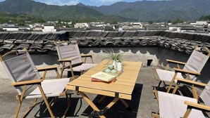 Une terrasse sur le toit, avec des toits traditionnels recouverts de tuiles, une table en bois avec des chaises, et une vue sur les montagnes en arrière-plan.