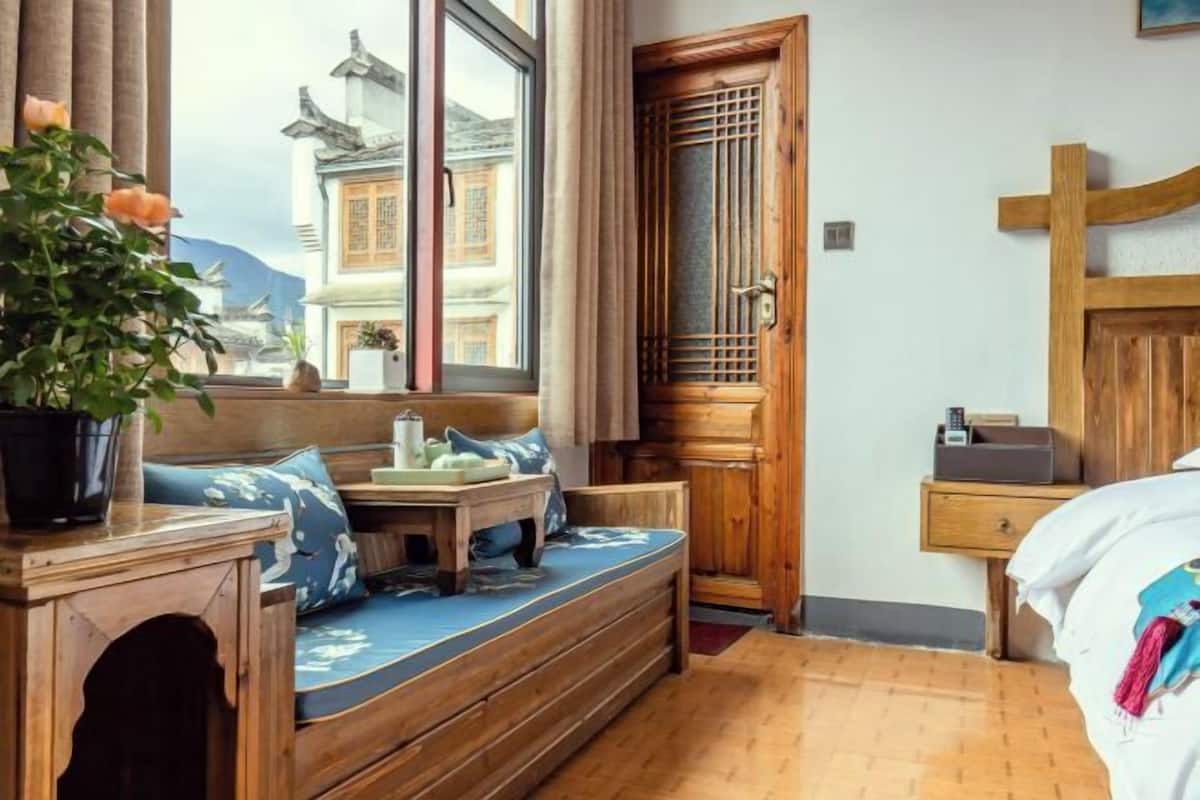 Une pièce comprenant un banc en bois, un lit, une table de chevet et une fenêtre donnant sur des bâtiments traditionnels.