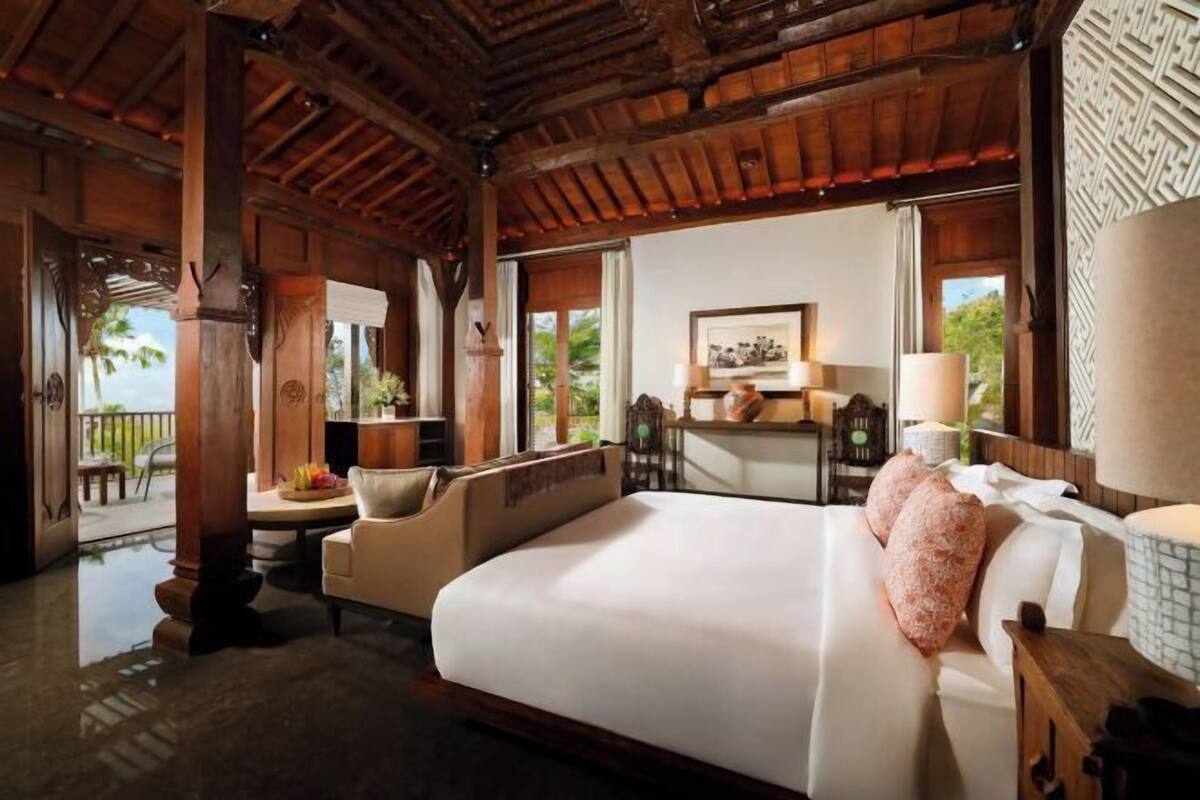 A spacious bedroom with a large bed, wooden ceiling beams, a desk with a chair, and a view of the outdoors.