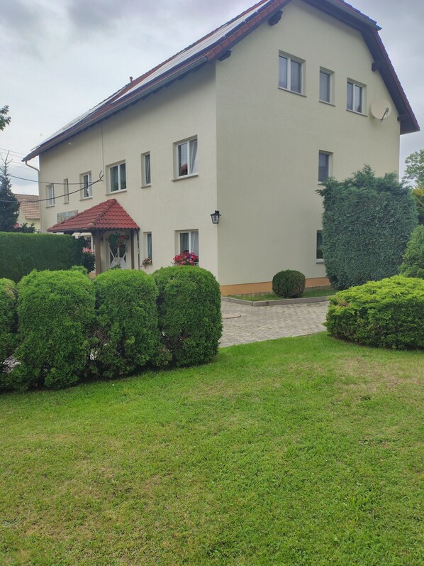 Ein zweistöckiges Gebäude mit rotem Ziegeldach, einer Satellitenschüssel und einem kleinen Garten mit geschnittenen Büschen und einem gepflasterten Weg.