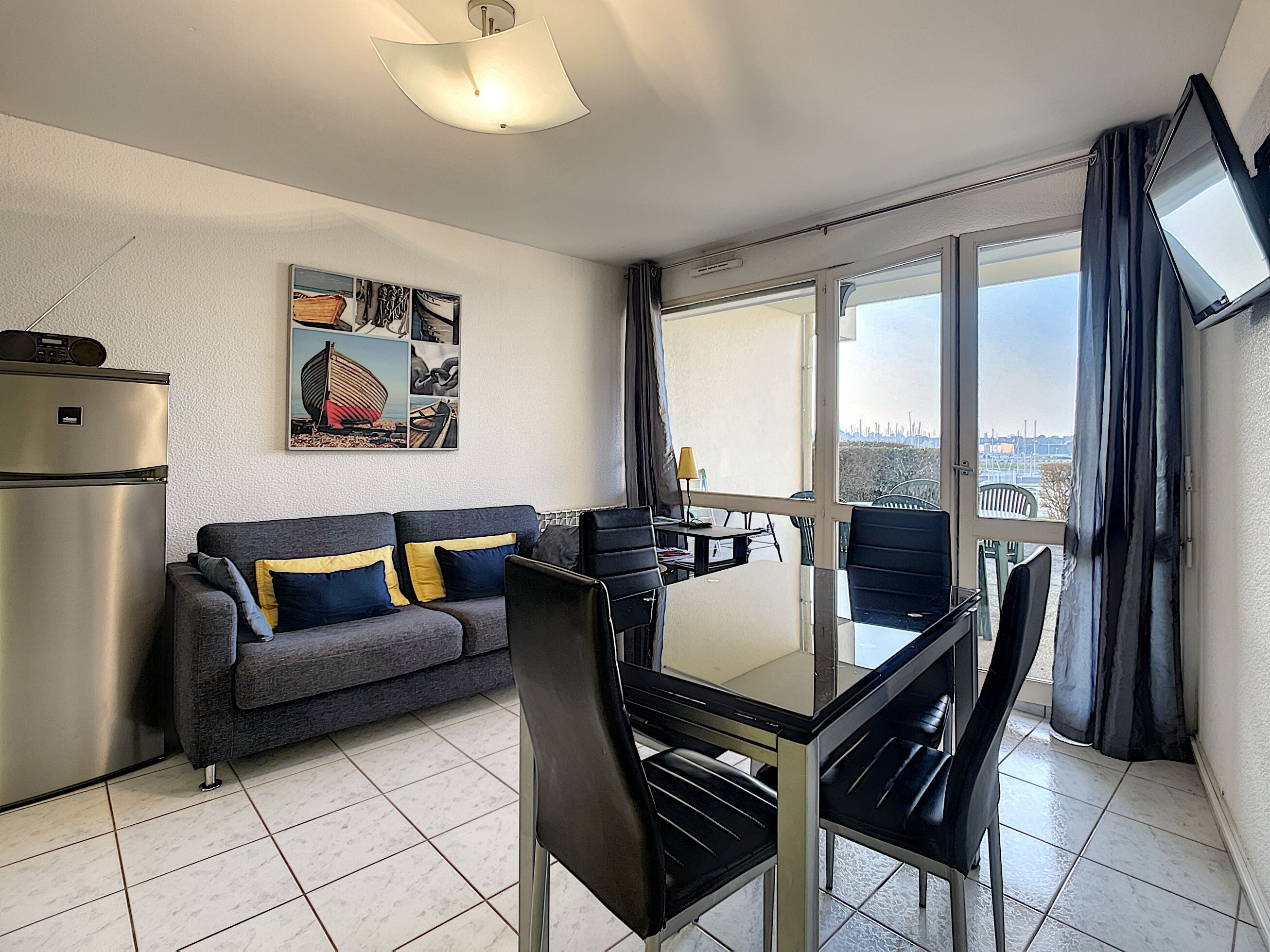 A modern living room with a sofa, dining table, chairs, a refrigerator, and a large window with a city view.