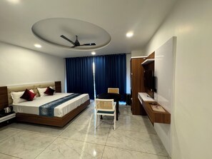 A modern hotel room with a bed, a desk, a chair, a ceiling fan, and a TV mounted on the wall. - Basant Haveli (Roorkee)