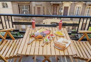 Une table en bois, sur laquelle sont disposés divers aliments et boissons, est placée sur un balcon offrant une vue sur les bâtiments.