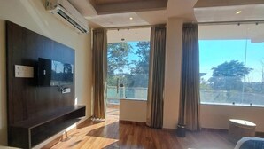 A spacious room with a large window, a flat-screen TV mounted on a dark wood panel, and a wooden floor. - Castle Inn Homestay  (Dehradun)