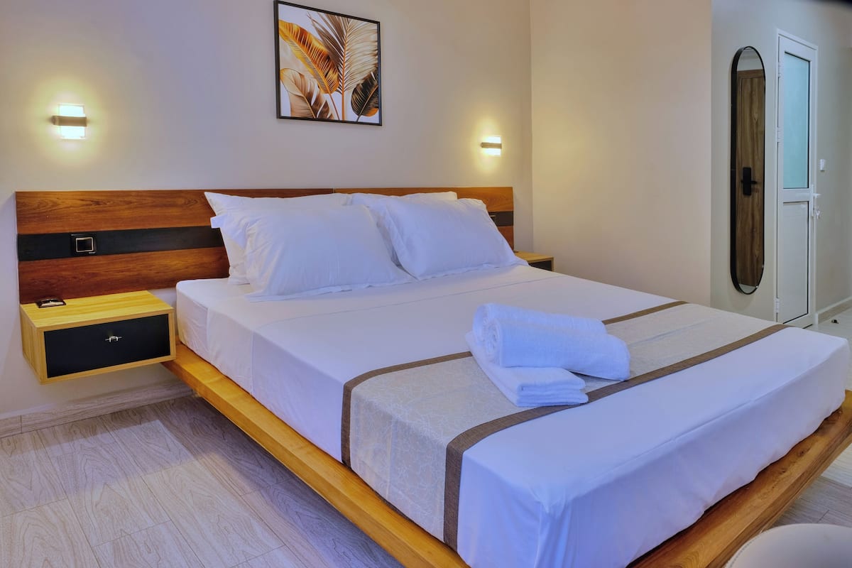 Una cama bien hecha con sábanas blancas y una toalla doblada, una mesita de noche de madera con cajón, y una lámpara de pared.