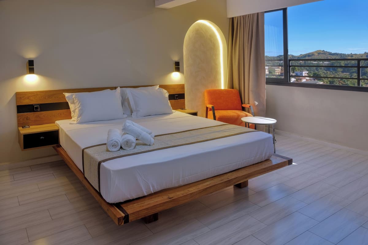 Una habitación de hotel moderna con una cama grande, un cabecero de madera, dos mesitas de noche, una pequeña mesa redonda y vistas de la ciudad a través de un ventanal amplio.