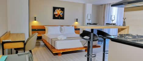 A compact hotel room with a bed, a desk, a chair, a small kitchen area, and a dining table.
