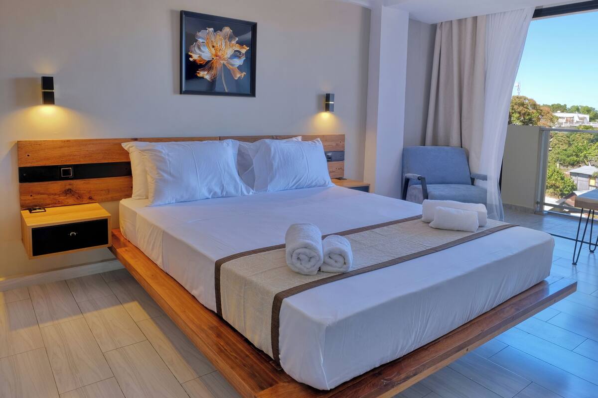 Habitación de hotel moderna con una cama grande, dos mesitas de noche, una silla y vistas al exterior.