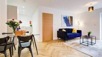 Una sala moderna con piso de espiga, un sofá azul oscuro con cojines amarillos, una mesa de comedor de vidrio y sillas, y una mesita auxiliar con una planta.