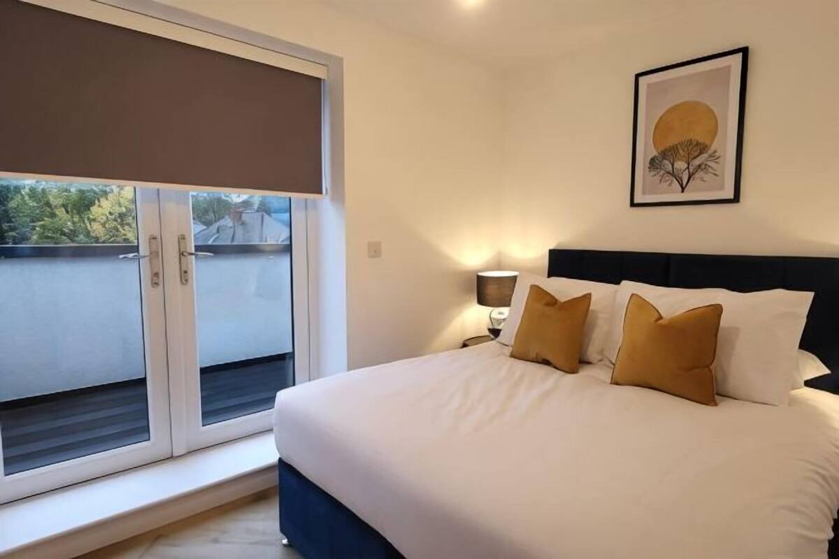 Une chambre à coucher avec un lit, une fenêtre avec un store, une lampe fixée au mur et un tableau encadré.