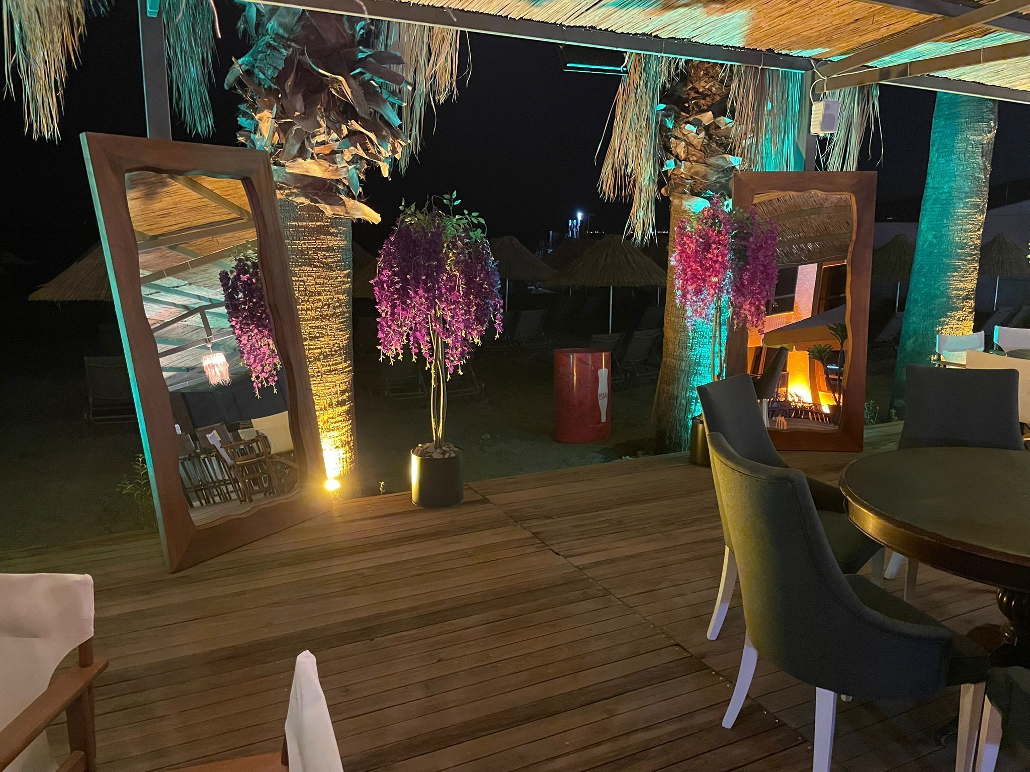 An outdoor seating area with wooden flooring, chairs, and tables. There are decorative plants and a fire feature.