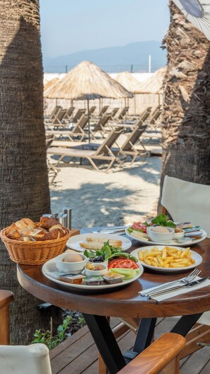 A table set with a variety of dishes including bread, salad, and fries, overlooking a beach with lounge chairs and umbrellas. - Grand Cennet Beach Hotel (Edremit)