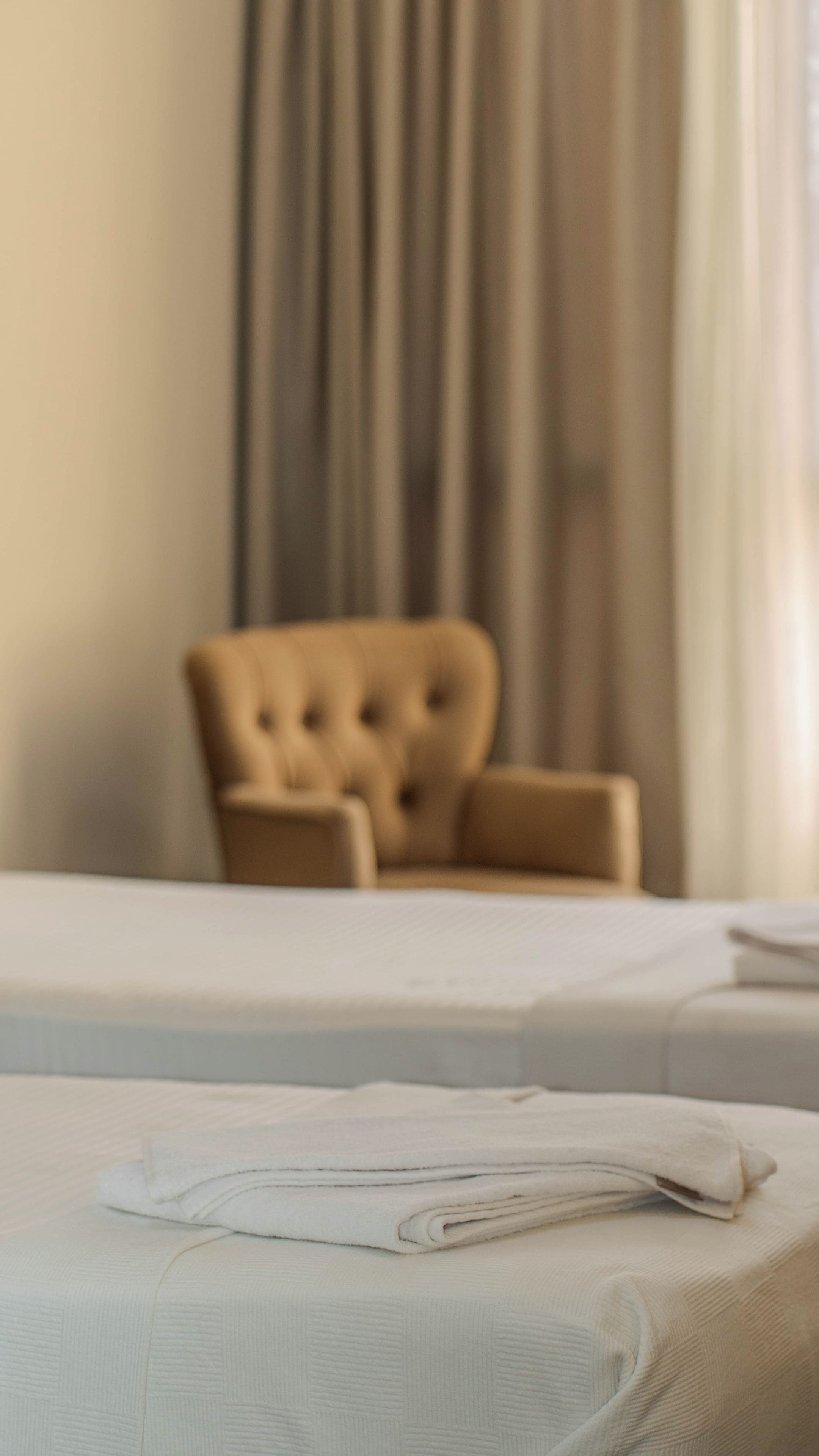 Een bed met een netjes opgemaakt wit deken en een gestoffeerde fauteuil op de achtergrond.