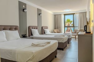 Habitación de hotel con tres camas, un escritorio y vistas a palmeras.