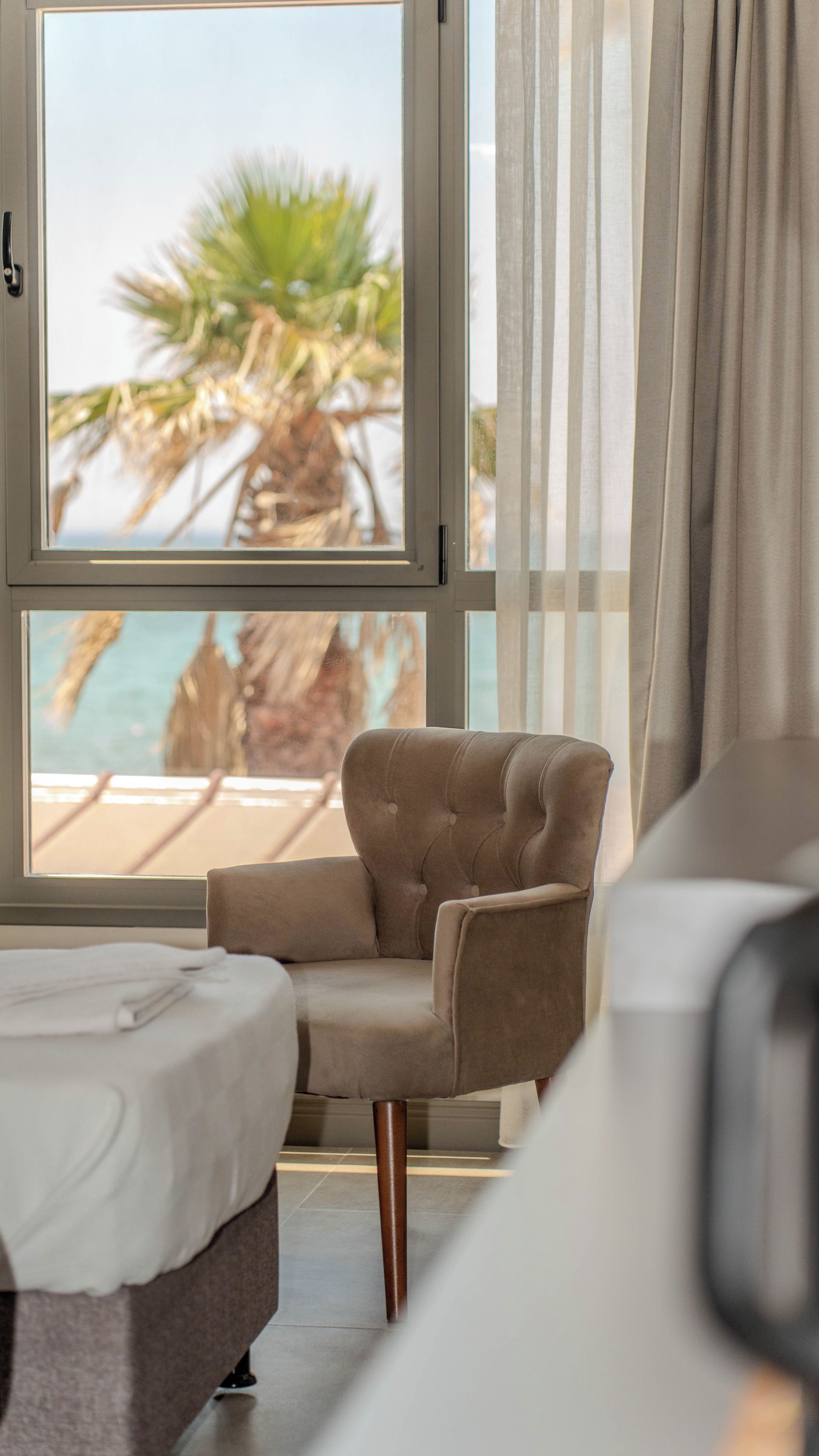 A hotel room with a bed, a chair, a view of the beach, and curtains.
