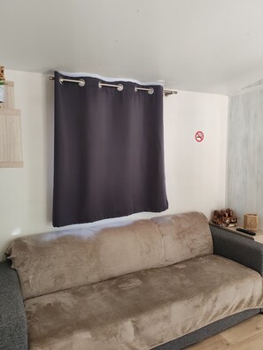 A room with a beige sofa, a dark curtain, a no-smoking sign, and a small wooden shelf with items. - Attractive, fully equipped mobile home for 4 people   (Samoëns)