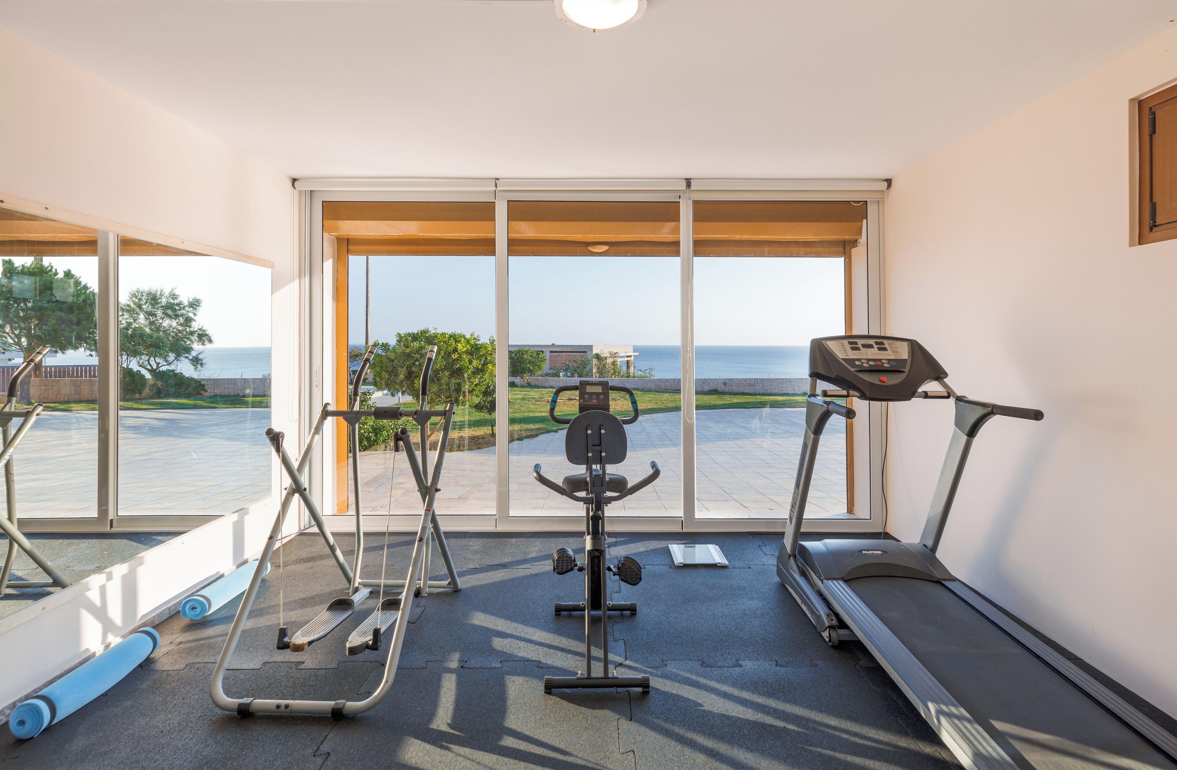 Un gimnasio con cintas de correr, elípticas y pesas, con vista al mar y un edificio al fondo.