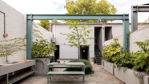 A stone-paved patio with a green table and benches, surrounded by plants and a white building with a garage door. - Chic Seddon Retreat Near the CBD (Seddon)