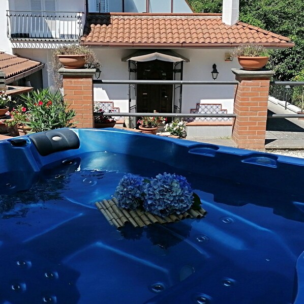 Un bain à remous agrémenté d’une composition florale décorative et un espace au bord de la piscine avec des plantes en pot, le tout avec un bâtiment en arrière-plan.