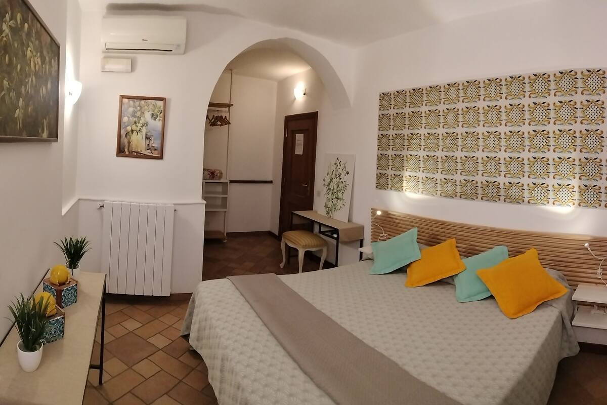 A hotel room with a bed, a desk, a chair, a radiator, a potted plant, and a wall with a decorative pattern.