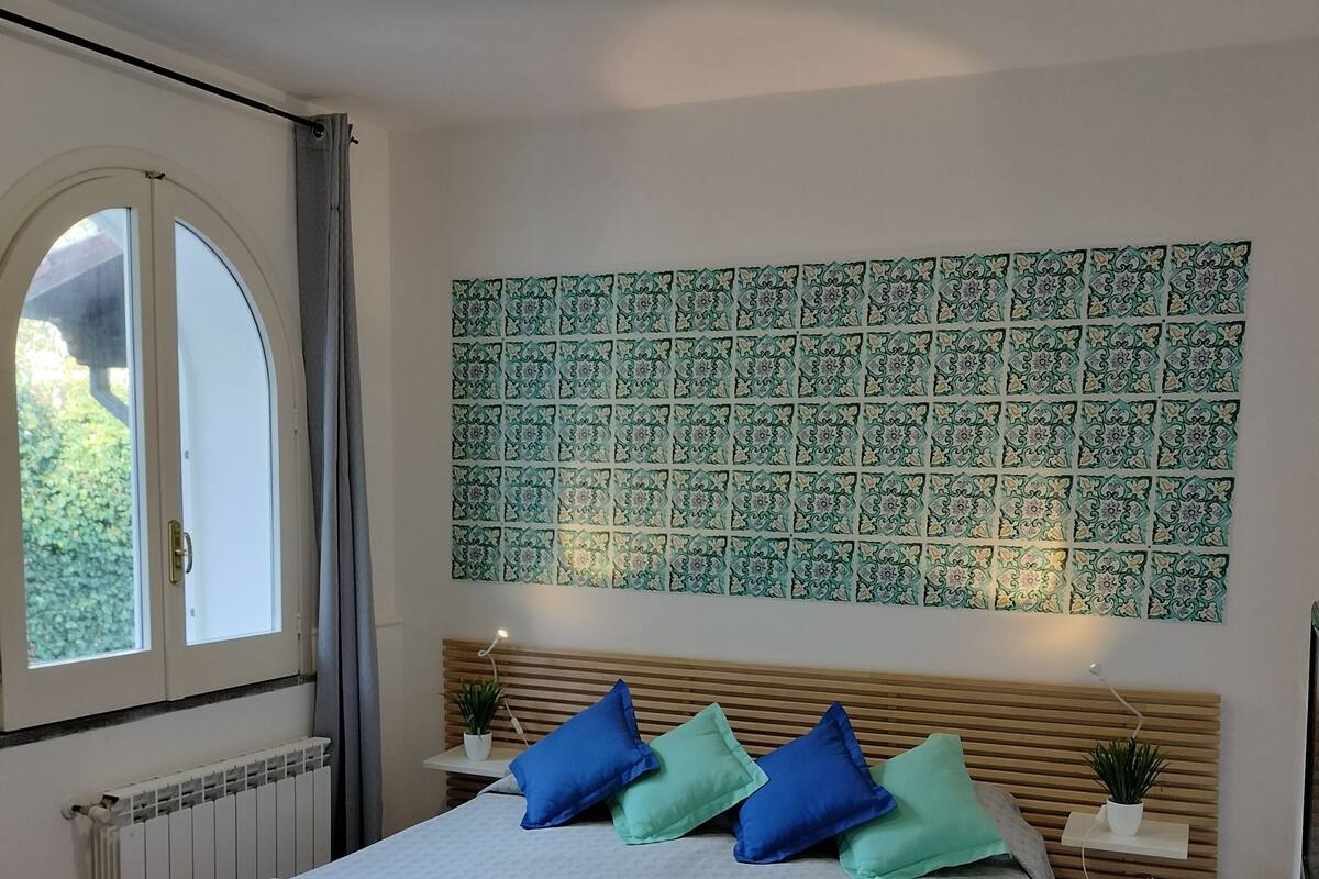 A bedroom with a bed, a window, a radiator, a wall tapestry, and bedside tables with plants.