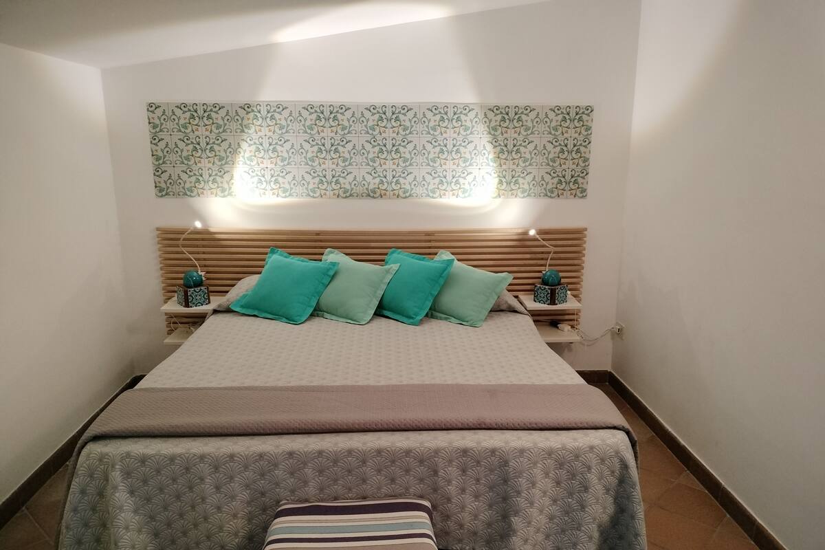 A neatly arranged bedroom with a bed, pillows, a bedside table, and a wall-mounted light fixture.