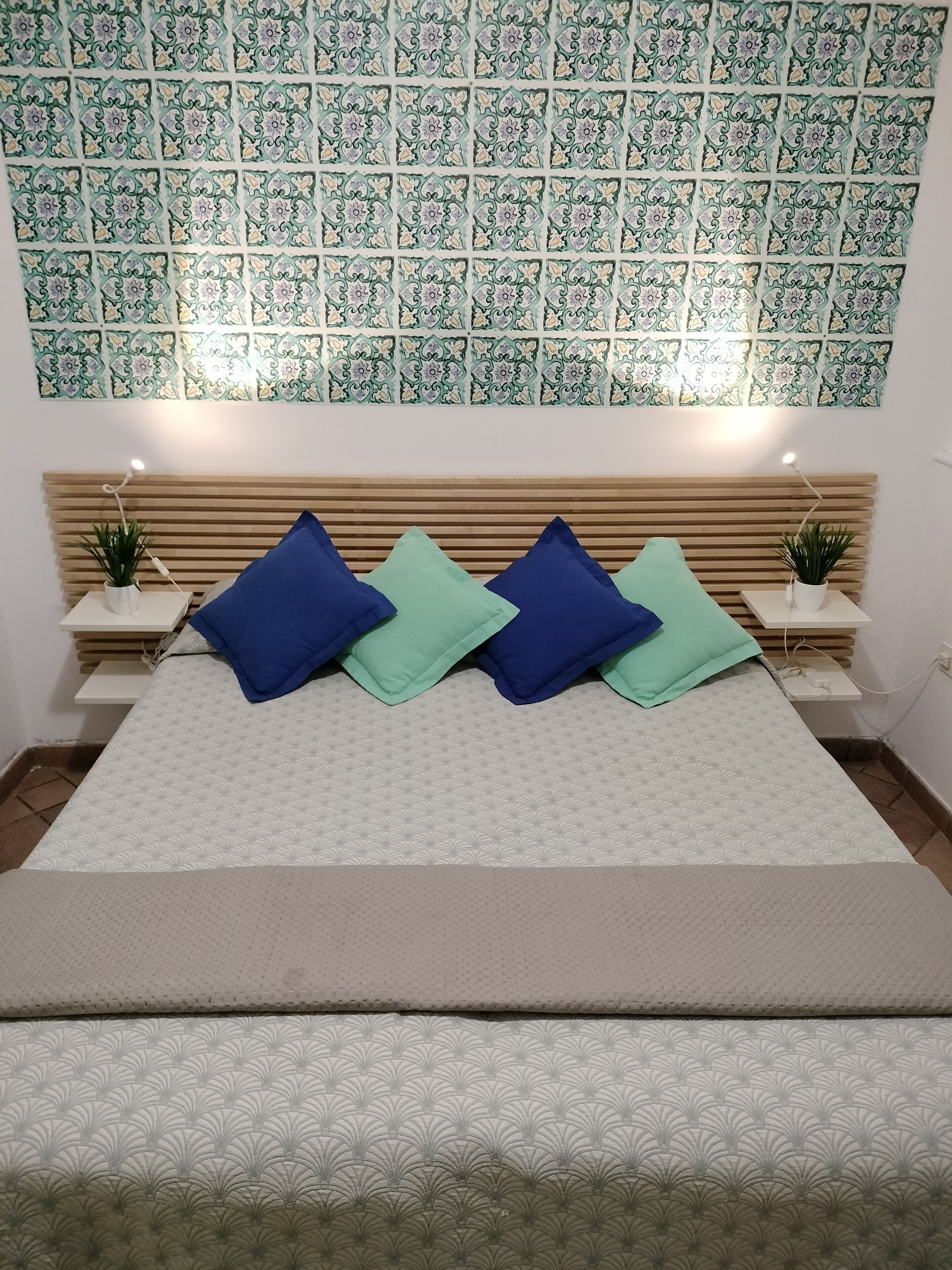 Un dormitorio ordenado con una pared estampada, una cama con cubrecama color beige y dos mesitas de noche con lámparas y plantas.