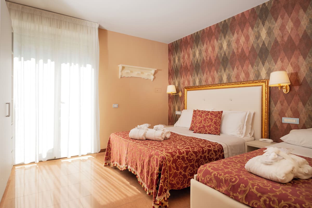 A hotel room with two beds, a large window with sheer curtains, a patterned wall, and a small shelf above the beds.