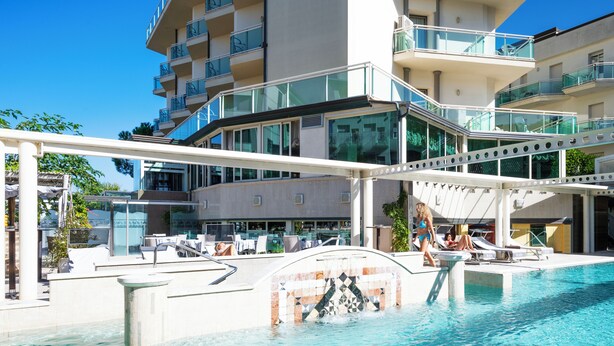 A swimming pool with a glass balustrade, lounge chairs, and a modern building with balconies in the background. - Hotel Lady Mary (Milano Marittima)