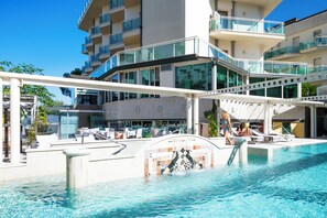 A swimming pool with a glass balustrade, lounge chairs, and a modern building with balconies in the background. - Hotel Lady Mary (Milano Marittima)