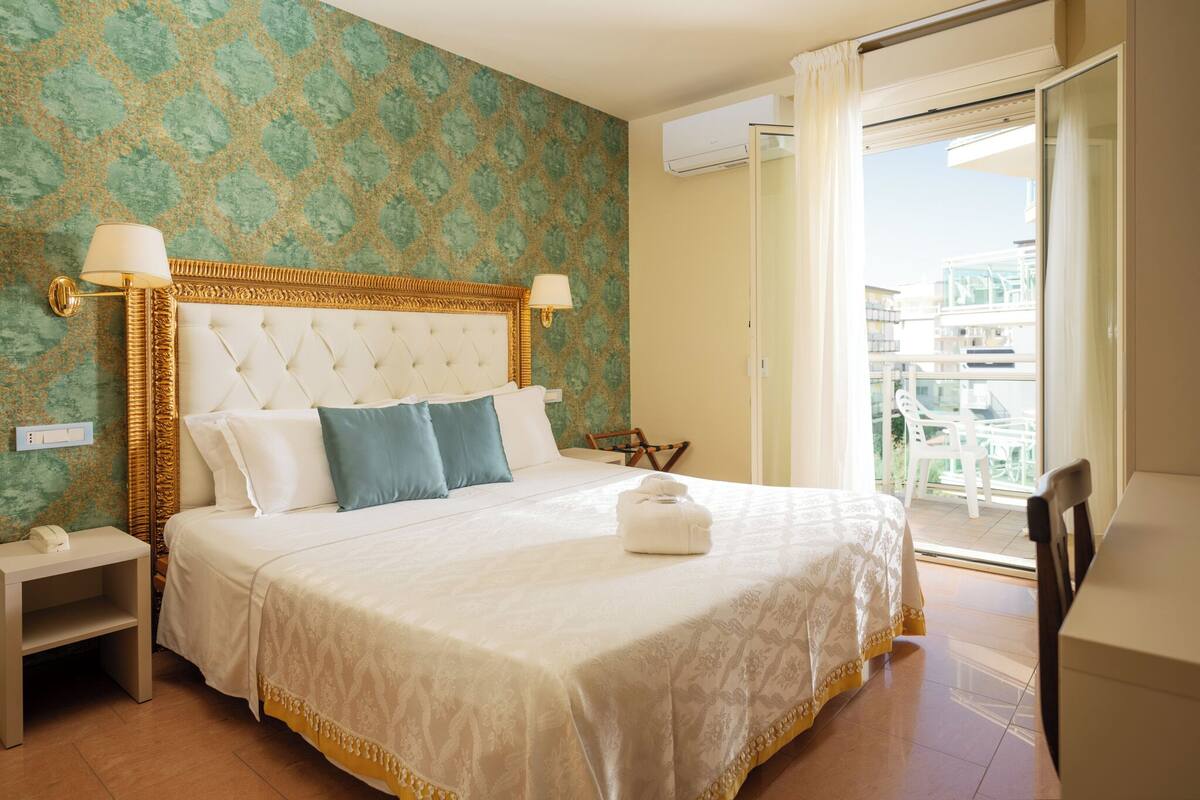 A hotel room with a large bed, two bedside lamps, a nightstand, a balcony with a chair, and a view of buildings.
