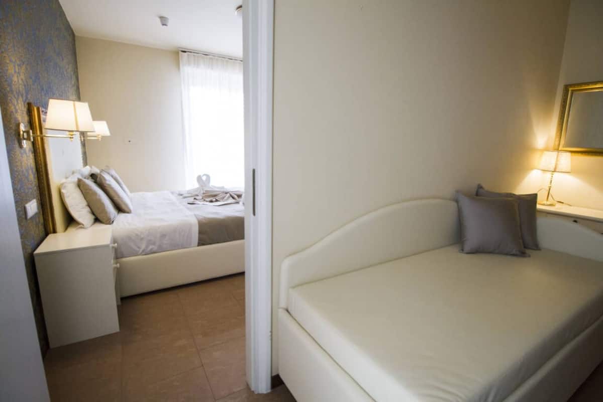 A hotel room with a bed, bedside table, lamp, and a view of another room.