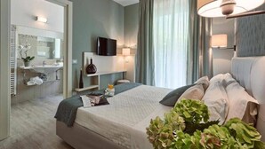 A modern hotel room with a bed, a TV, a desk with a lamp, a bathroom with a sink and mirror, and a large window with curtains. - Hotel Lady Mary (Milano Marittima)