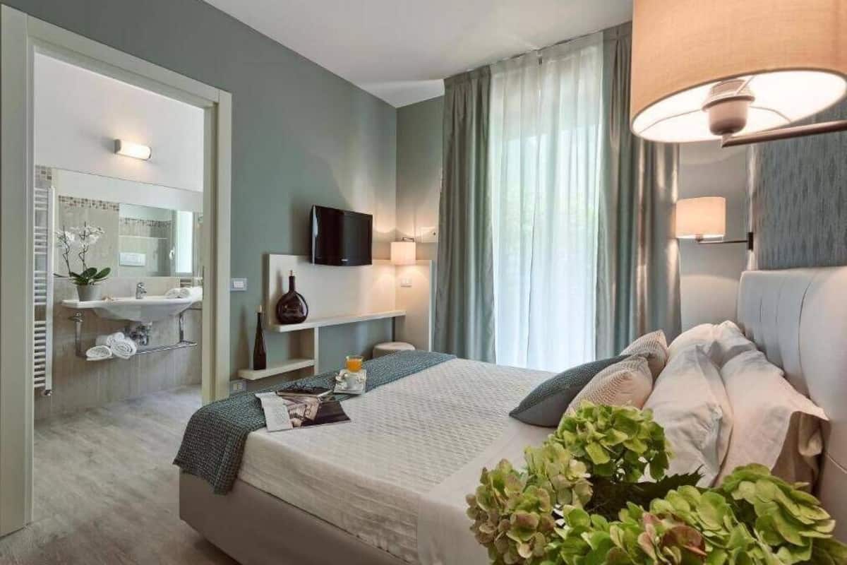 A modern hotel room with a bed, a TV, a desk with a lamp, a bathroom with a sink and mirror, and a large window with curtains.
