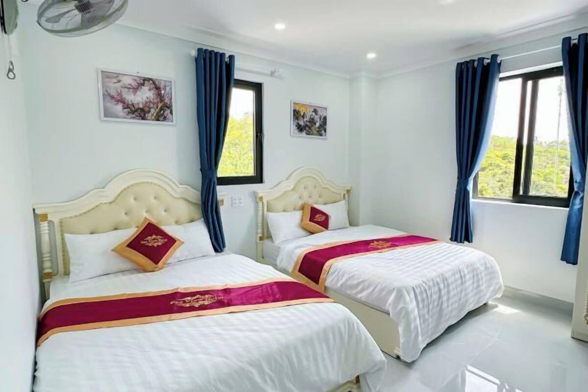 A hotel room with two beds, each with a white headboard and a red and gold bedspread. There are two windows with blue curtains, and the walls are painted white.