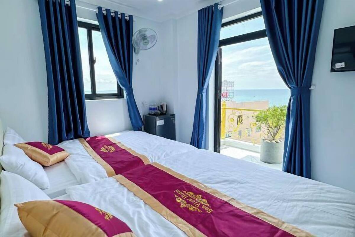 A hotel room with a bed, blue curtains, a window, a fan, and a view of the city.