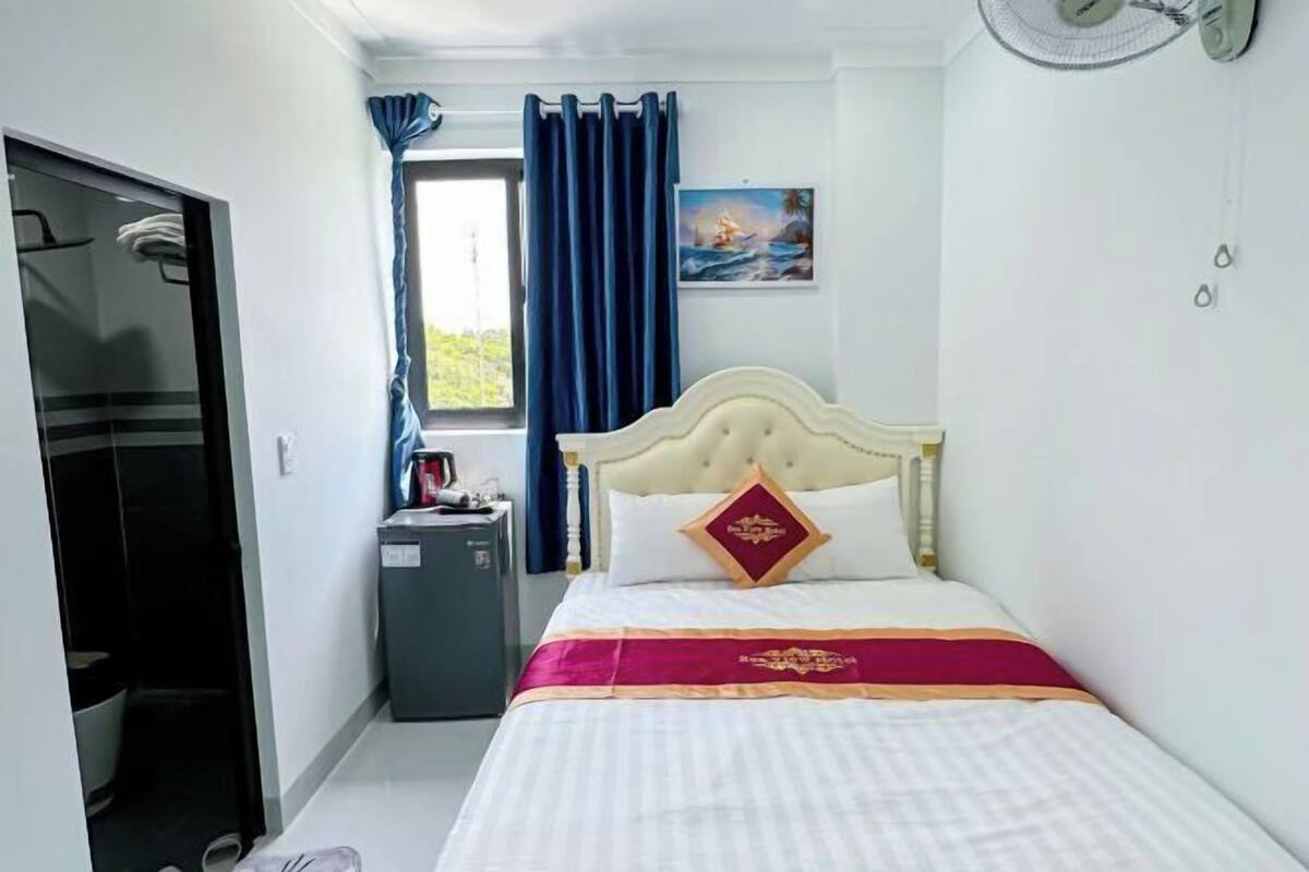 A bedroom with a bed, a window with curtains, a small desk, and a fan.