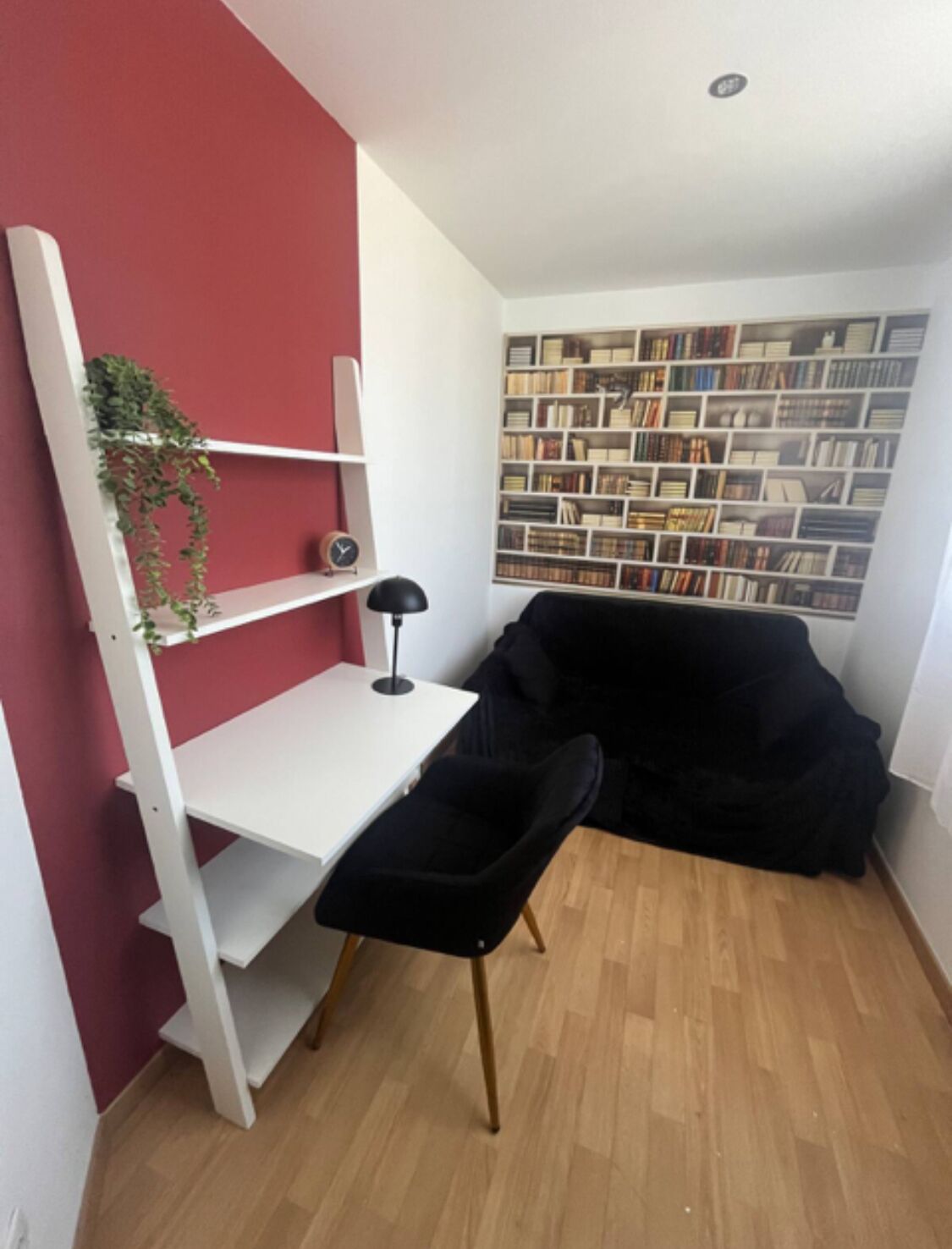 A compact room with a white desk, a black chair, a bookshelf, and a red accent wall.