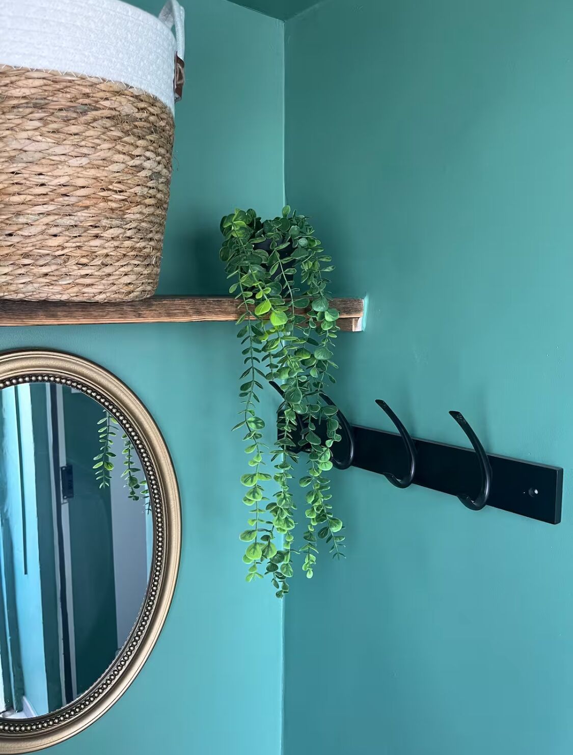 Een teal wand met een geweven mand, een hangplant, een spiegel en een zwart wandhaak.